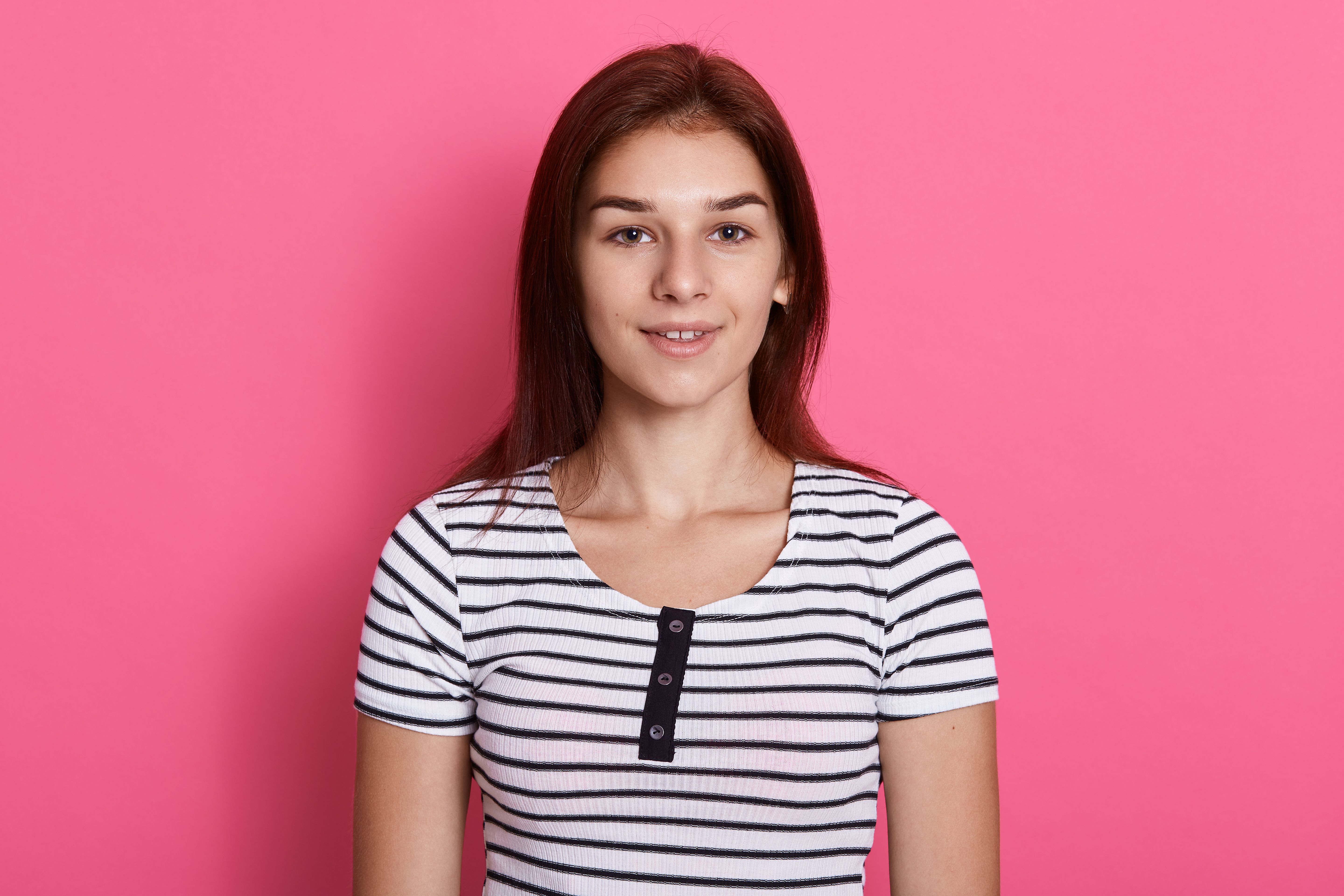 young attractive teenage girl wearing striped casual t shirt posing against pink wall with charming smile looks happy